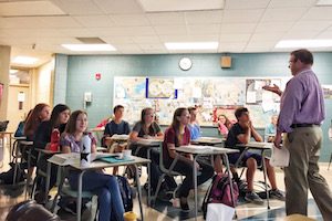 The image features a classroom of teenagers sitting at their desk while a teacher talks in front of them.