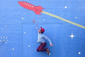 This image features a young boy jumping in the air, in front of a blue wall with a red rocketship painted on it.