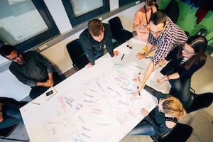 This image features a group of 6 people collaborating on a large piece of white paper on a table.